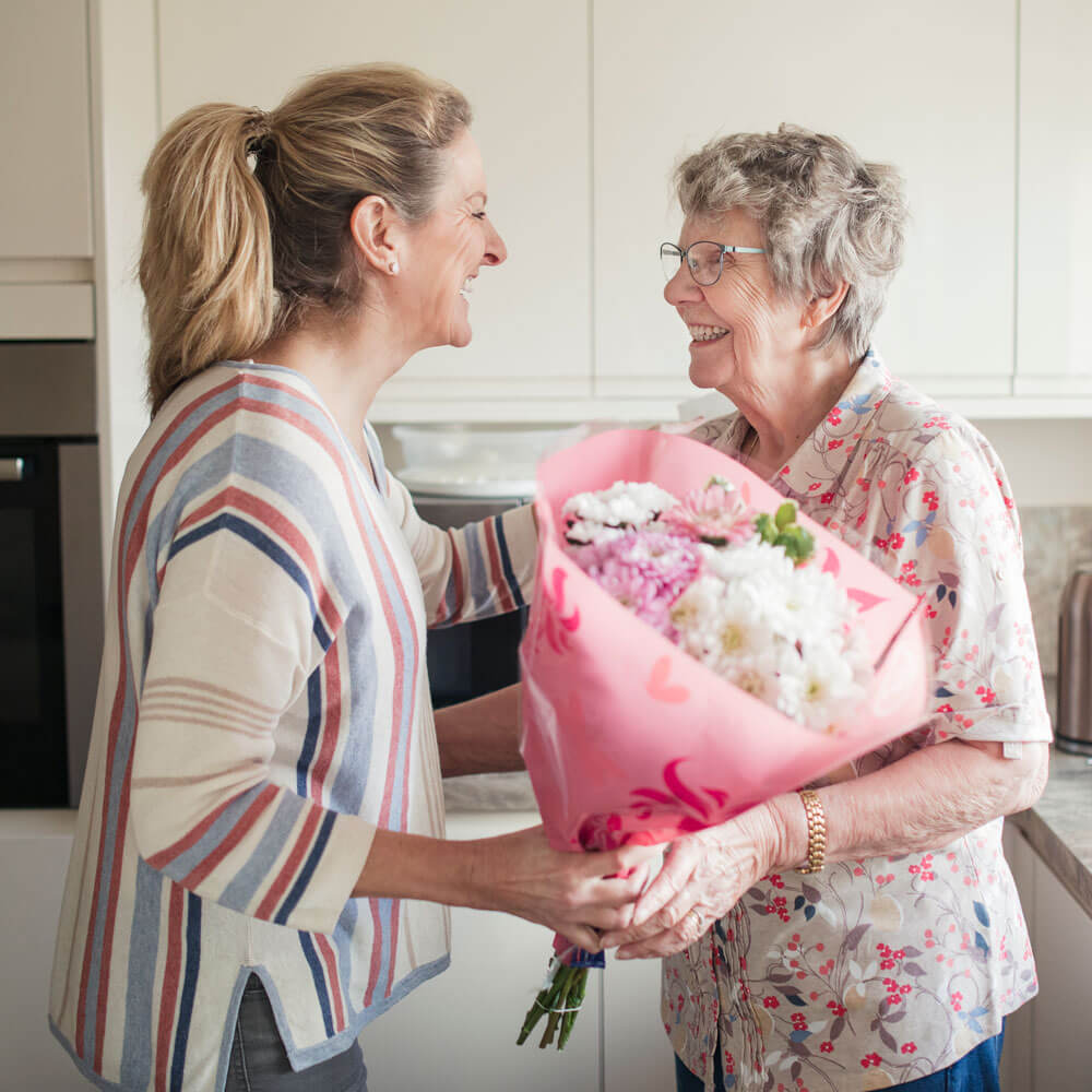 Daughter or caregiver standing in kitchen, handing a senior woman flowers